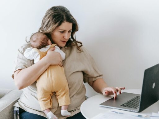 Concentrated young female freelancer embracing newborn while sitting at table and working remotely on laptop at home